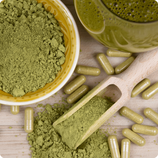 Overhead view of a wooden scoop filled with green kratom powder, surrounded by a yellow bowl of powder, scattered kratom capsules, and a glass of green liquid on a wooden surface.