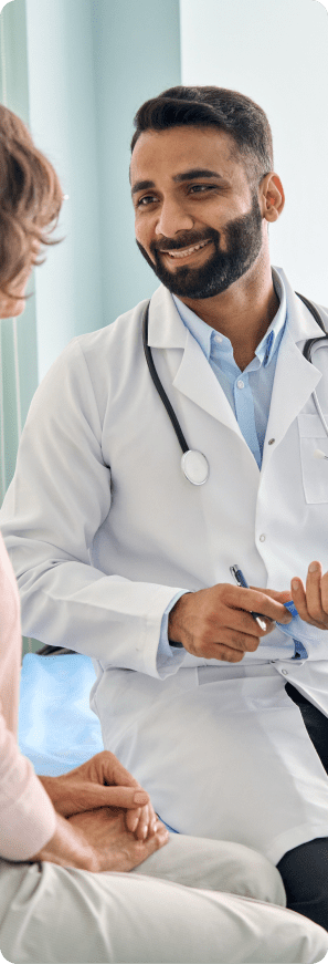 Doctor speaking with a seated patient during a consultation, smiling and holding a pen in a clinical office setting