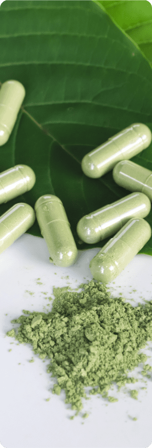 Close-up view of green kratom capsules resting on a large green leaf, next to a small pile of loose green kratom powder on a white surface.