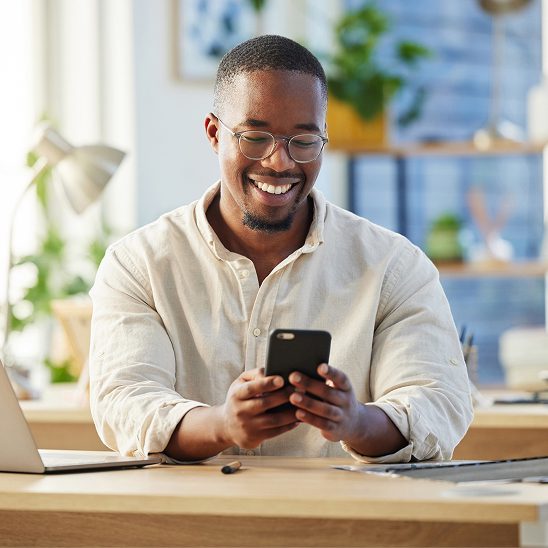 Man sitting at desk smiling while looking at smartphone, with office in the background