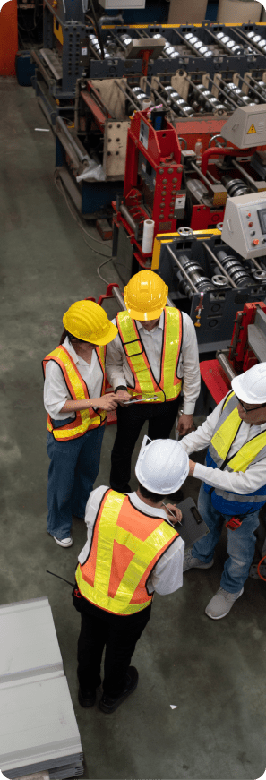 Manufacturing professionals in safety vests review project details on a tablet inside an industrial facility.