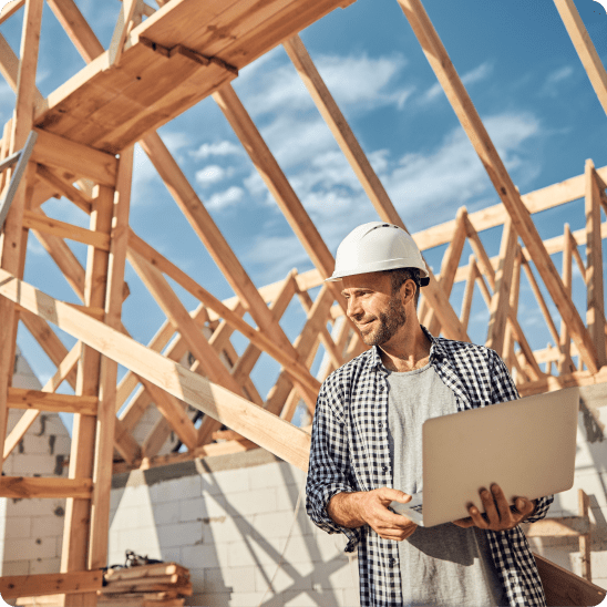 Builder in a hard hat reviews plans on a laptop at a residential construction site.