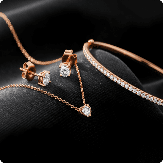 Close-up of rose gold jewelry with diamond earrings, a bracelet, and a heart pendant arranged on black fabric.