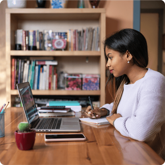 Student taking notes while attending an online course on a laptop at home