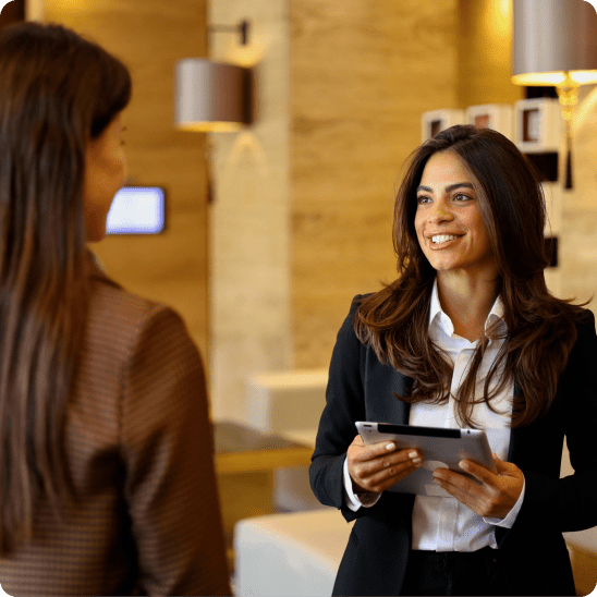 Hospitality professional greets a guest while holding a tablet in a hotel lobby.